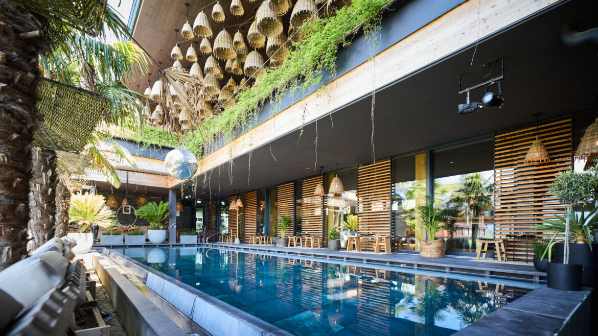 Modernes Freibad mit Holzdekor, hängenden Weidenlampen und üppigem Grün an einem sonnigen Tag. (vergrößerte Ansicht)