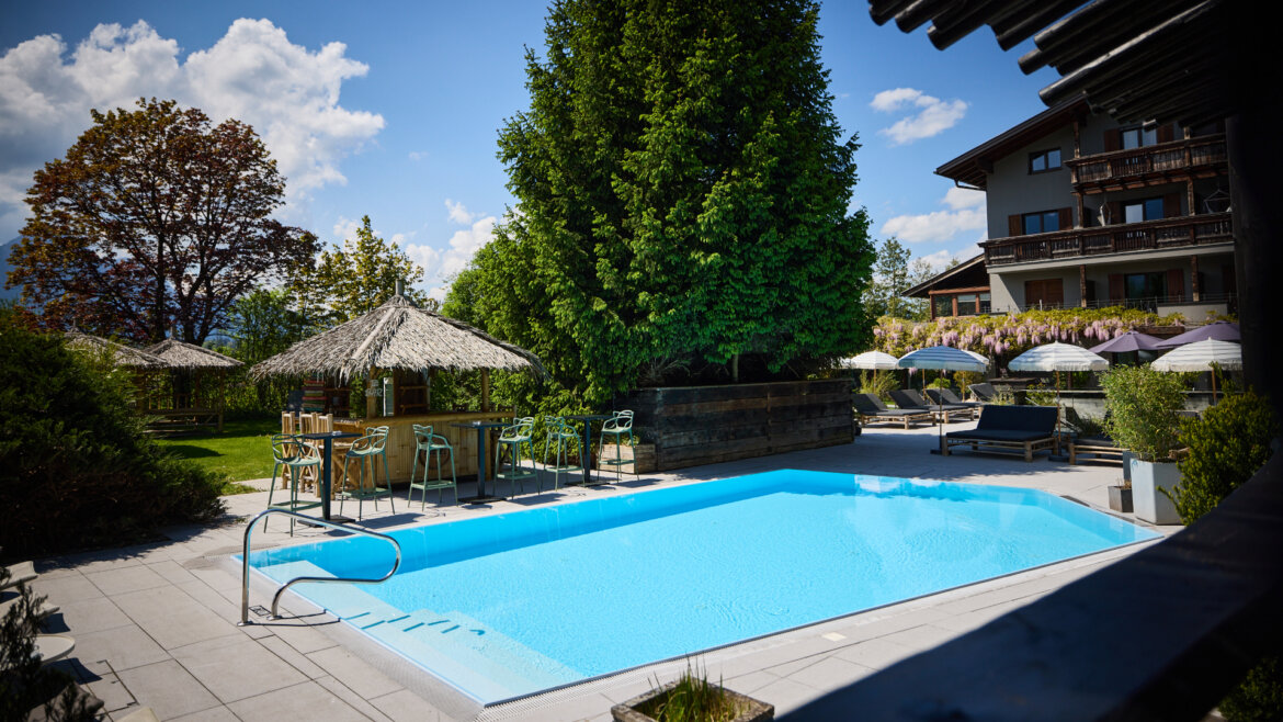 Swimmingpool im Freien, umgeben von Bäumen, Liegestühlen und Sonnenschirmen an einem sonnigen Tag. (vergrößerte Ansicht)