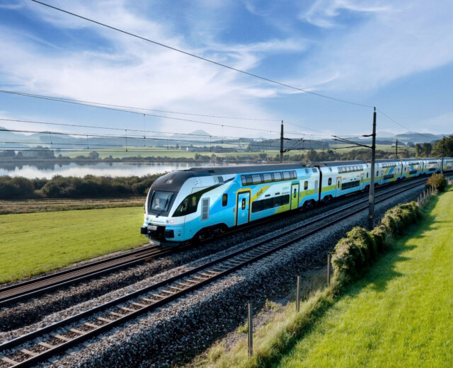 WESTbahn_fruehling Ein moderner Doppelstockzug fährt auf Schienen durch eine grüne Landschaft unter blauem Himmel.