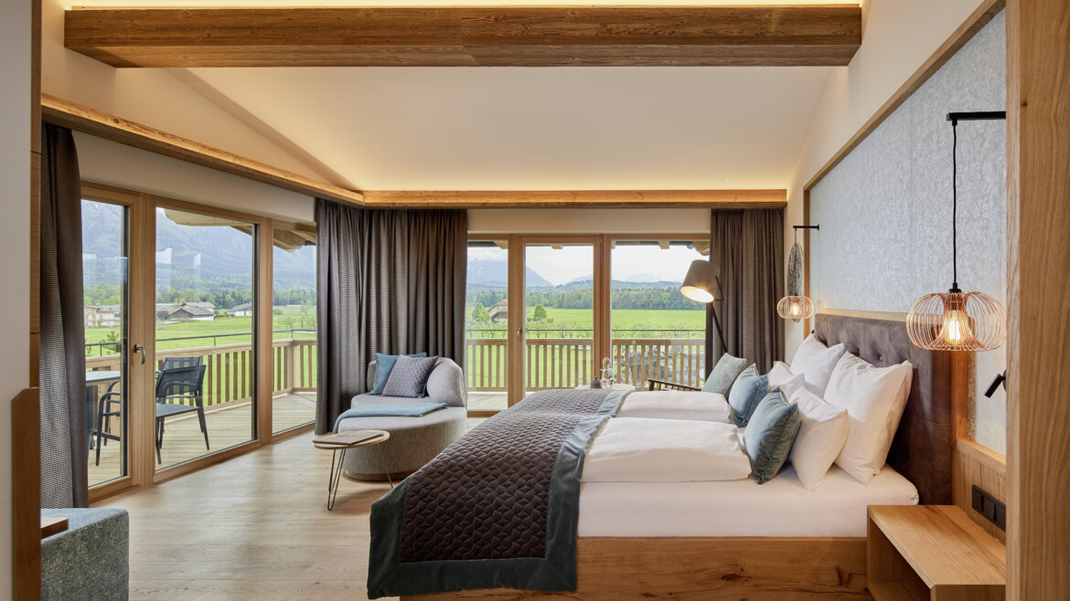 Geräumiges, modernes Schlafzimmer mit großen Fenstern, Doppelbett, Balkon und Blick auf grüne Felder und Berge. (vergrößerte Ansicht)
