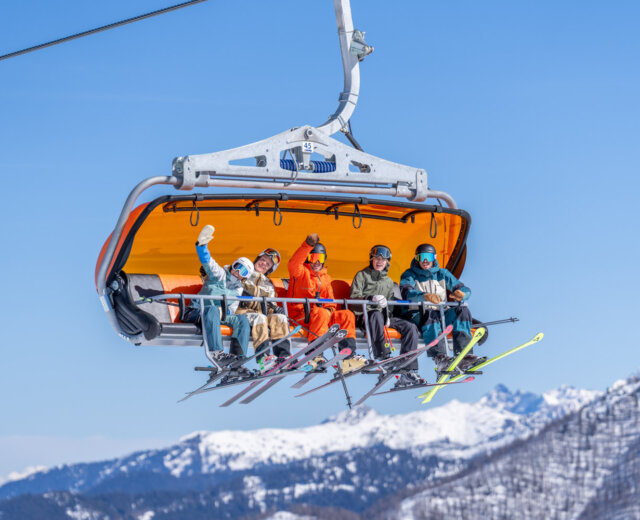 8er Sessellift Flachau (c) Flachau Tourismus – Christian Lorenz Fünf Skifahrer sitzen lächelnd und winkend in einem Skilift, die orangene Bubble oben, mit verschneiten Bergen im Hintergrund.