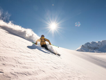 Skifahrer in gelber Jacke beim Skifahren auf Neuschnee mit strahlendem Sonnenschein und klarem blauen Himmel.