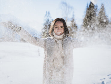 Chillout Schneebegeisterung © SalzburgerLand Tourismus, Branislav Rohal, Lächelnde Person in Winterkleidung wirft Schnee in die Luft in einer verschneiten, sonnenbeschienenen Landschaft mit Bäumen.