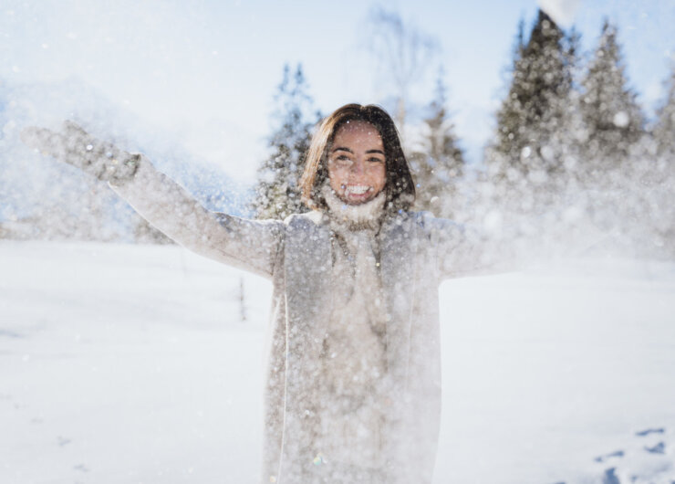 Lächelnde Person in Winterkleidung wirft Schnee in die Luft in einer verschneiten, sonnenbeschienenen Landschaft mit Bäumen.