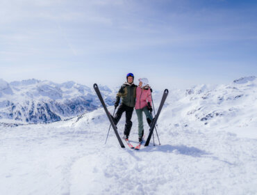 Zwei Personen in Skiausrüstung stehen dicht beieinander auf einem verschneiten Berg, die Skier vor ihnen gekreuzt.