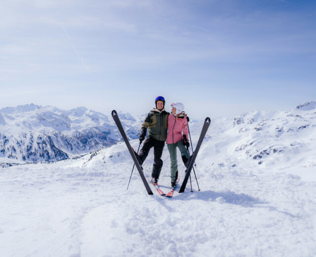 Zwei Personen in Skiausrüstung stehen dicht beieinander auf einem verschneiten Berg, die Skier vor ihnen gekreuzt.