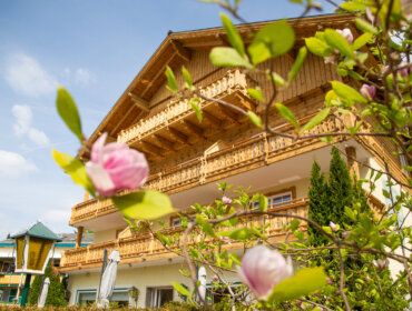 Ein hölzernes Gebäude im Chalet-Stil mit Balkonen, die durch blühende rosa Blumen und grüne Blätter zu sehen sind.