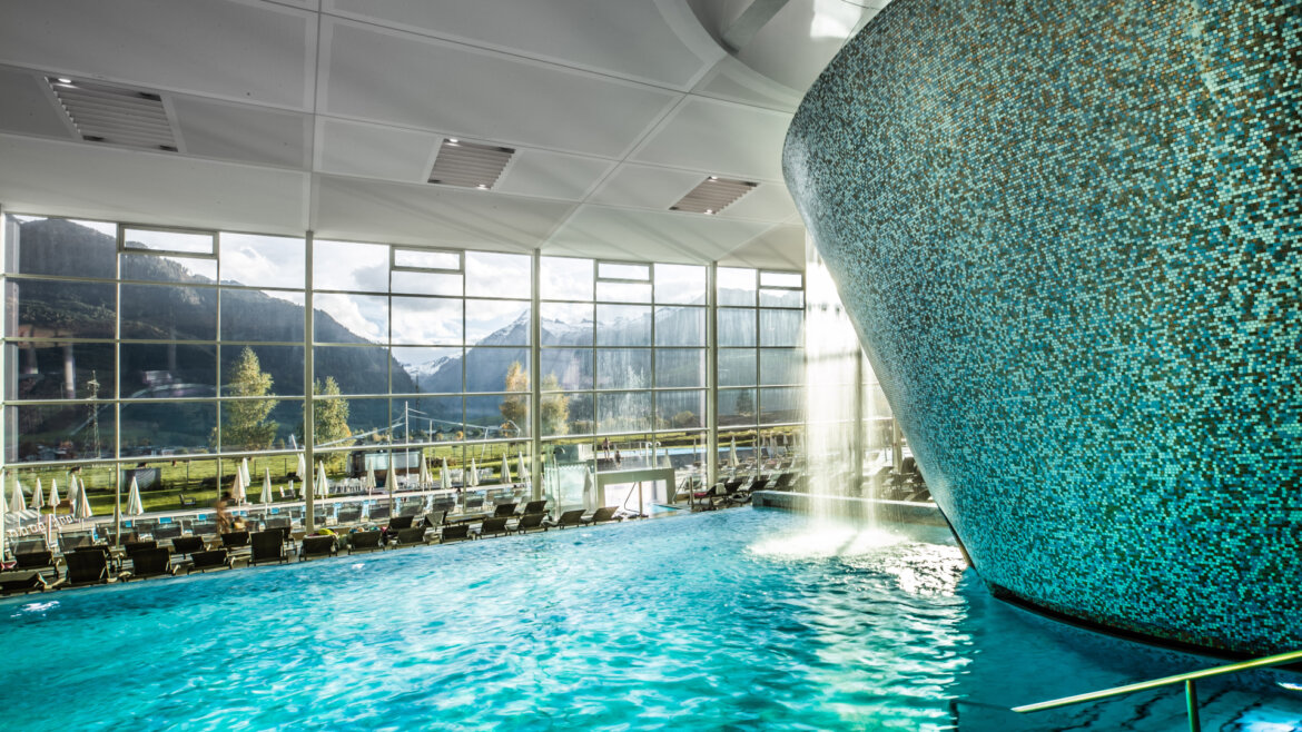 Innenpool mit Mosaikwand und Wasserfall, große Fenster mit Blick auf die Berge und aufgereihte Liegestühle. (vergrößerte Ansicht)