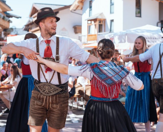 Menschen in traditioneller österreichischer Kleidung tanzen im Freien bei einer festlichen Veranstaltung mit Schaulustigen im Hintergrund.
