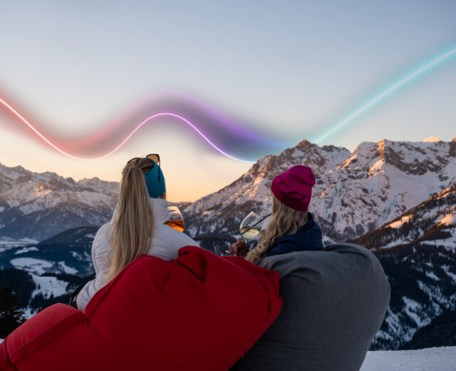 March Glow3 Abendstimmung (c) HochkoenigTourismusGmbH Zwei Frauen entspannen sich auf Sitzsäcken in verschneiten Bergen bei Sonnenuntergang, bunte Lichtspuren am Himmel über ihnen.