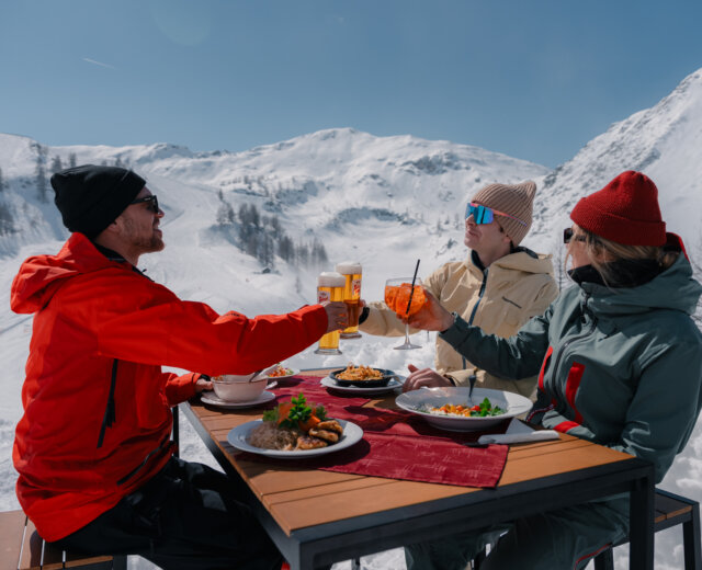 Zauchensee Kulinarischer Genuss beim Skifahren (c) roastmedia_zauchensee Drei Personen in Winterjacken stoßen mit Getränken an einem Tisch mit Essen an, umgeben von verschneiten Bergen und schönsten sonnigen Wetter.