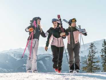 Saalbach Skidamen © Christoph Johann, Saalbach Drei gutgelaunte Damen mit Skiern auf dem Schultern an einem sonnigen Tag. Berge im Hintergrund.
