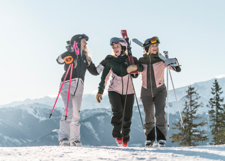 Drei gutgelaunte Damen mit Skiern auf dem Schultern an einem sonnigen Tag. Berge im Hintergrund.
