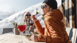 Saalbach Hüttenkulinarik © Christoph Johann Zwei Personen in Winterjacken essen und trinken im Freien an einem sonnigen, verschneiten Ort in den Bergen.