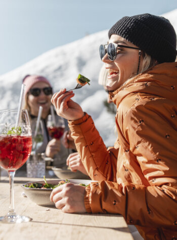 Zwei Personen in Winterjacken essen und trinken im Freien an einem sonnigen, verschneiten Ort in den Bergen.