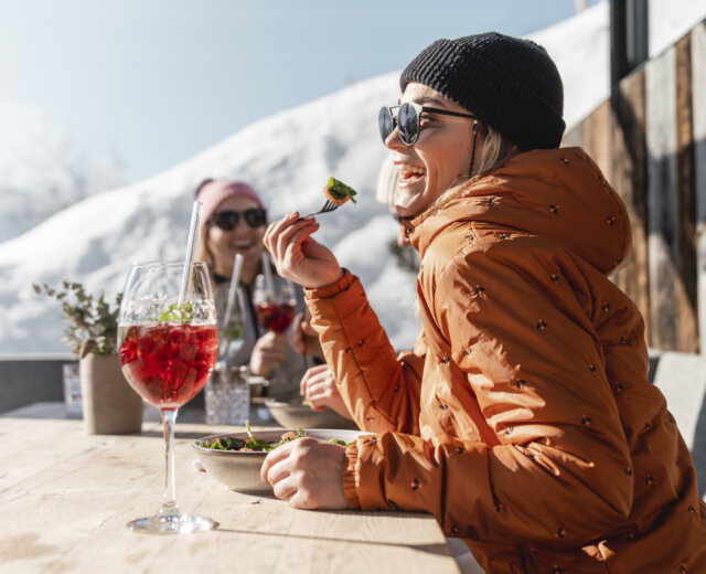 Zwei Personen in Winterjacken essen und trinken im Freien an einem sonnigen, verschneiten Ort in den Bergen.