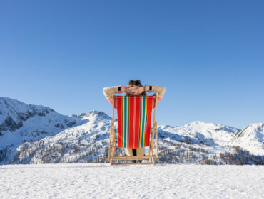 Person, die sich auf einem bunten Liegestuhl im Schnee entspannt, mit Blick auf verschneite Berge unter einem klaren blauen Himmel.