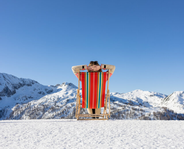 Person, die sich auf einem bunten Liegestuhl im Schnee entspannt, mit Blick auf verschneite Berge unter einem klaren blauen Himmel.
