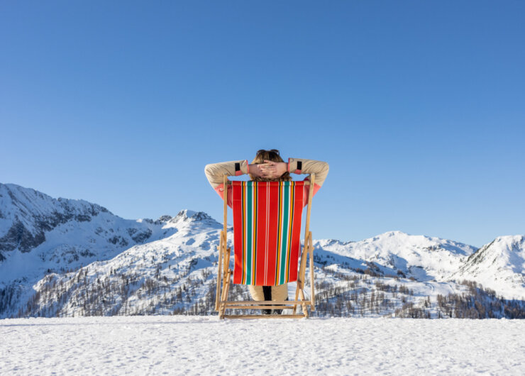 Winter Chillout Liegestuhl, (c) SalzburgerLand Tourismus, Hansi Heckmair Person, die sich auf einem bunten Liegestuhl im Schnee entspannt, mit Blick auf verschneite Berge unter einem klaren blauen Himmel.