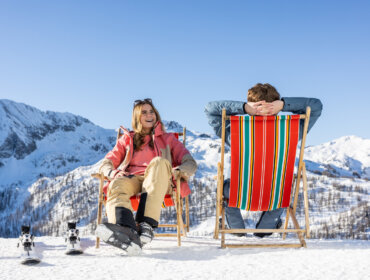 Winter Chillout, (c) SalzburgerLand Tourismus,1679 Eine Frau und ein Mann genießen ein Winterchillout in Liegestühlen auf Schnee, Berge und Skiausrüstung in der Nähe.