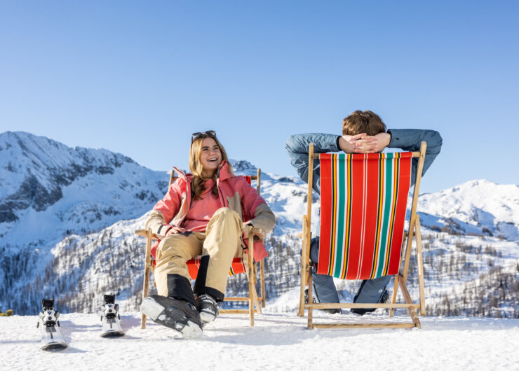 Eine Frau und ein Mann genießen ein Winterchillout in Liegestühlen auf Schnee, Berge und Skiausrüstung in der Nähe.