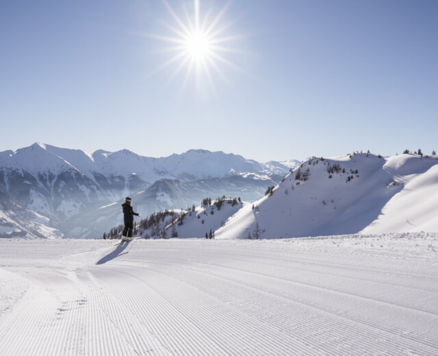 Skifahren Rauris (c) Ferienregion Nationalpark Hohe Tauern – Branislav Rohai Ein Skifahrer gleitet über eine sonnige, frisch präparierte Piste mit verschneiten Bergen im Hintergrund.