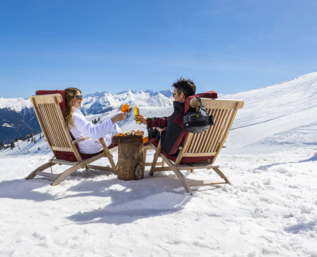 Zwei Personen in Skikleidung stoßen in Liegestühlen auf einem verschneiten Berg mit Skiern und schneebedeckten Gipfeln im Hintergrund an.