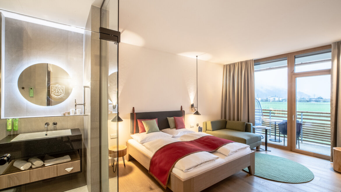 Helles, modernes Hotelzimmer mit Doppelbett, Sofa, Balkon, Glasdusche und Blick auf die grünen Felder draußen. (vergrößerte Ansicht)