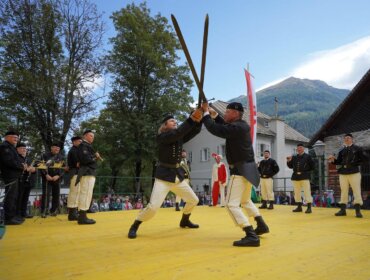 Zwei Männer in traditioneller Kleidung fechten mit Holzschwertern auf einer Freilichtbühne, beobachtet von Musikern und einer Menschenmenge.