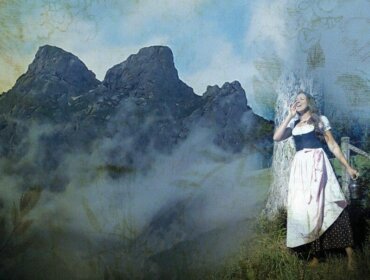 Eine Frau in traditioneller österreichischer Tracht steht lächelnd an einem Baum, mit nebligen Bergen im Hintergrund.