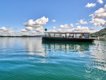 Ein kleines Ausflugsboot schwimmt auf einem ruhigen See unter einem blauen Himmel mit vereinzelten Wolken.