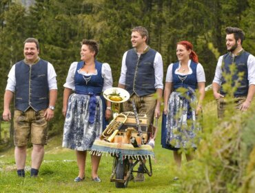 Fünf Personen in traditioneller österreichischer Kleidung stehen mit Musikinstrumenten in einer Schubkarre im Freien.