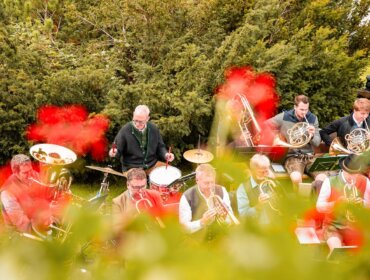 Eine Blaskapelle spielt unter freiem Himmel hinter roten Blumen und Grünpflanzen, wobei die Musiker verschiedene Instrumente spielen.
