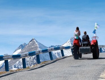 Ein Mann und eine Frau fahren mit einem Traktor eine Bergstraße hinauf, mit schneebedeckten Gipfeln im Hintergrund.