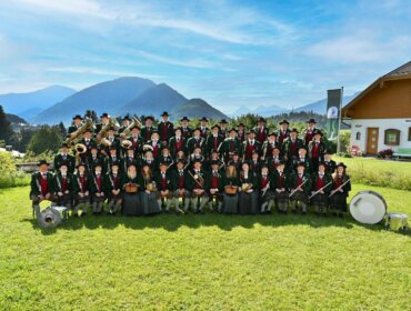 Ein großer Spielmannszug in traditioneller Kleidung posiert im Freien mit Instrumenten, im Hintergrund die Berge.