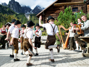 Jungen in traditioneller österreichischer Kleidung tanzen vor einer Blockhütte, während in der Nähe Musikanten Volksinstrumente spielen.