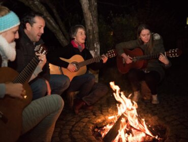 Vier Leute spielen Gitarren und singen gemeinsam am nächtlichen Lagerfeuer im Freien.