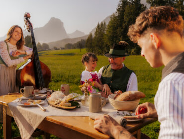 Vier Personen genießen eine Mahlzeit im Freien; eine Frau spielt einen Kontrabass, im Hintergrund sind die Berge zu sehen.