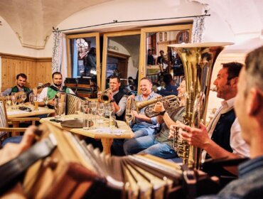 Eine Gruppe von Männern, die in einem gemütlichen, holzgetäfelten Raum gemeinsam Blas- und Streichinstrumente spielen.