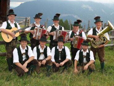 Neun Männer in traditioneller österreichischer Tracht mit Instrumenten, die im Freien vor Bergen posieren.