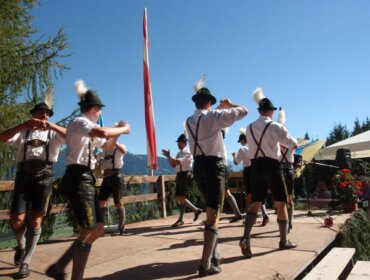 Männer in traditioneller österreichischer Tracht führen auf einer Holzbühne im Freien unter blauem Himmel einen Volkstanz auf.