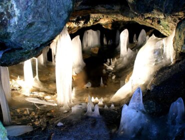 Aus dem Boden und der Decke der Höhle wachsen Eiszapfen und Eisgebilde, die von seitlichem Licht beleuchtet werden.