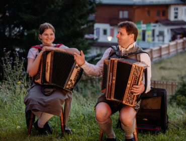 Zwei Personen in österreichischer Tracht spielen im Freien Akkordeon und lächeln sich in an.