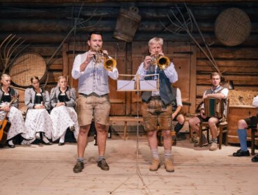 Zwei Männer in traditioneller österreichischer Kleidung spielen auf der Bühne Trompete, andere sitzen mit Instrumenten hinter ihnen.
