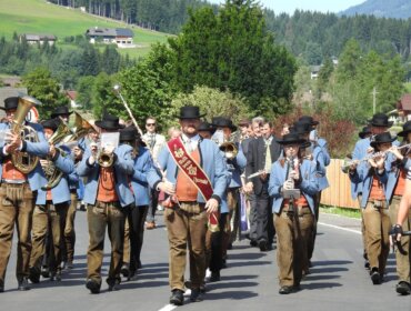 Eine Blaskapelle in österreichischer Tracht marschiert an einem sonnigen Tag im Freien in einem ländlichen Dorf auf.
