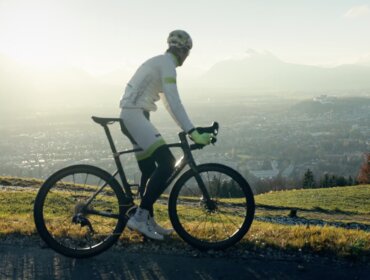 Radfahrer in weißer Kleidung fährt bei Sonnenaufgang auf einem Bergpfad mit Blick auf eine Stadt und ferne Berge.