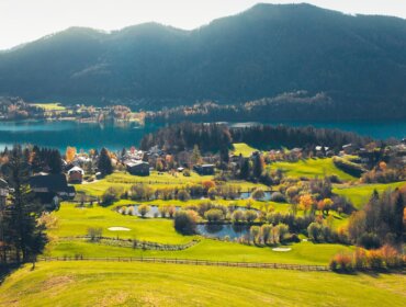 Ein malerischer Blick auf ein Dorf an einem See mit grünen Feldern, Bäumen und Bergen im Hintergrund.