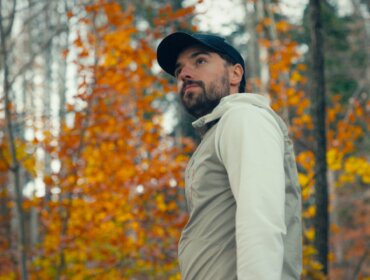 Herbstliche Auszeit am Fuschlsee | Mindful Escapes mit Max Bründl Mann in heller Jacke und Mütze steht in einem Wald mit Herbstblättern und orangefarbenem Laub im Hintergrund.