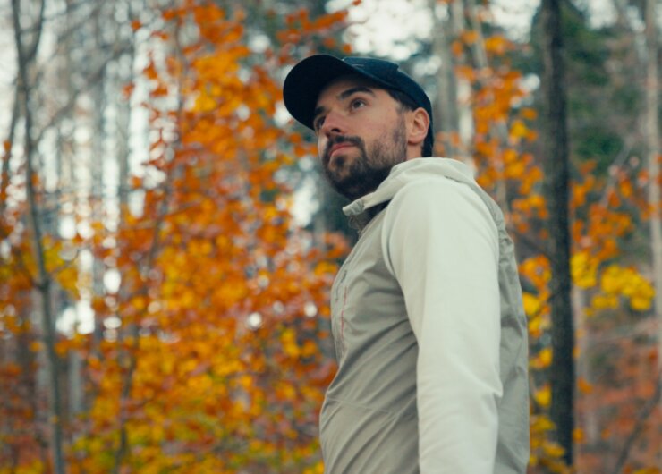 Mann in heller Jacke und Mütze steht in einem Wald mit Herbstblättern und orangefarbenem Laub im Hintergrund.