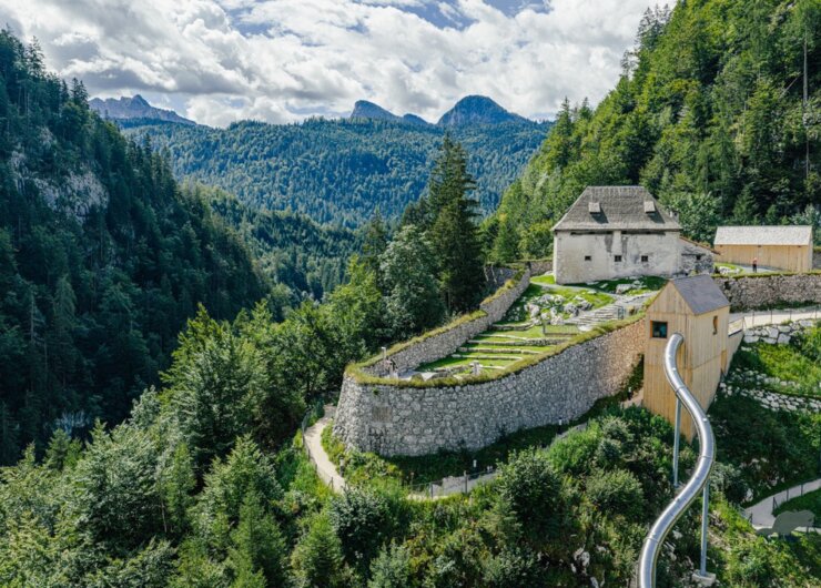 Ein Steingebäude mit terrassenförmig angelegten Gärten an einem bewaldeten Berghang, mit einer Metallrutsche und einer herrlichen Aussicht.